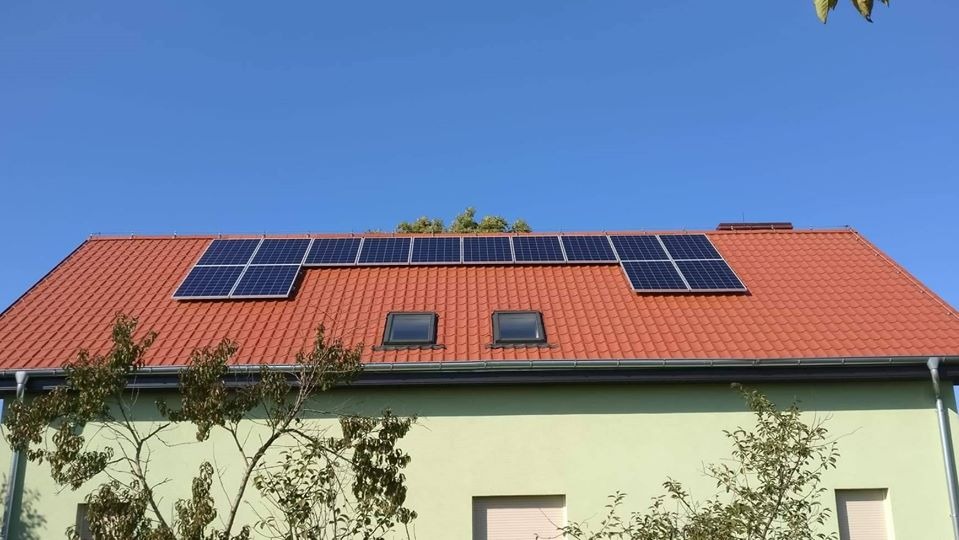 Panele fotowoltaiczne na dachu z czerwonej dachówki, dwa okna dachowe, jasna elewacja budynku, widok z dołu, błękitne niebo.