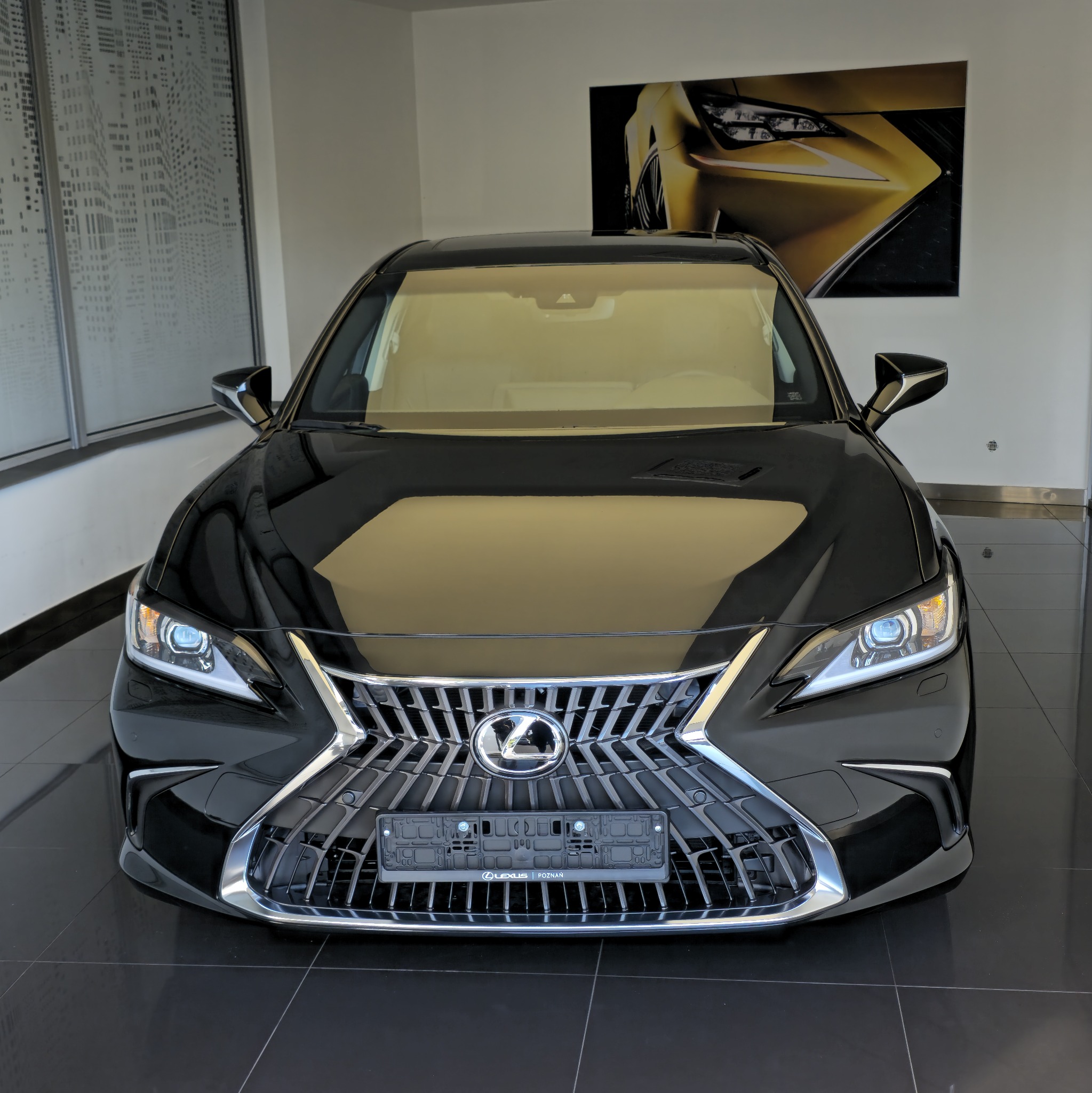 Czarny Lexus ES z chromowanym grillem, prezentowany w salonie samochodowym na tle grafiki auta i okna z motywem miejskim.