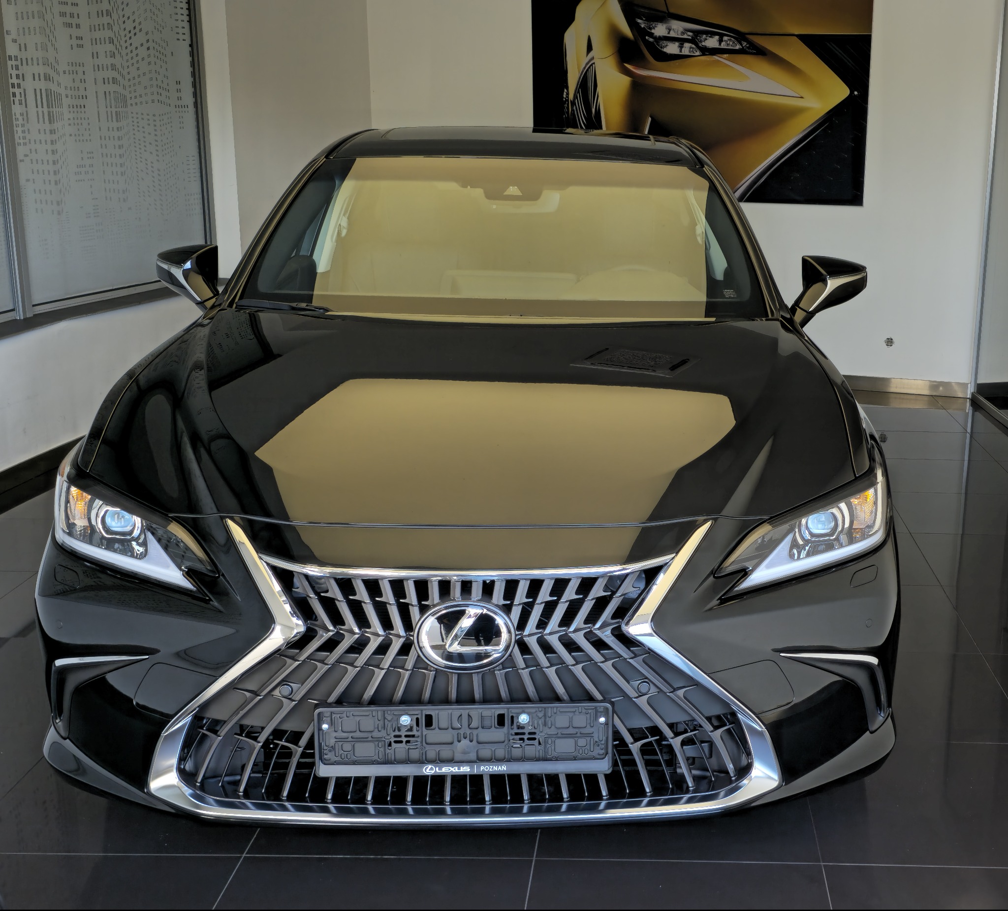 Czarny Lexus ES z chromowanym grillem na tle salonu samochodowego z grafiką innego modelu Lexusa. Widok z przodu, oświetlenie salonowe.