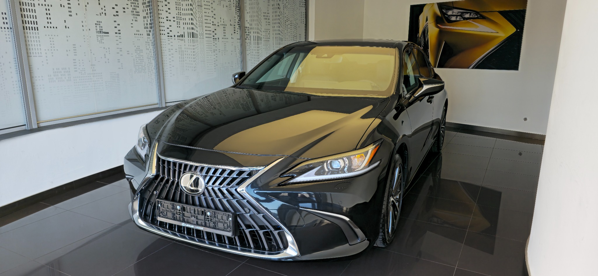 Czarny Lexus ES, elegancki sedan z chromowanym grillem na tle minimalistycznego wnętrza salonu samochodowego. Widoczny plakat z fragmentem innego Lexusa.