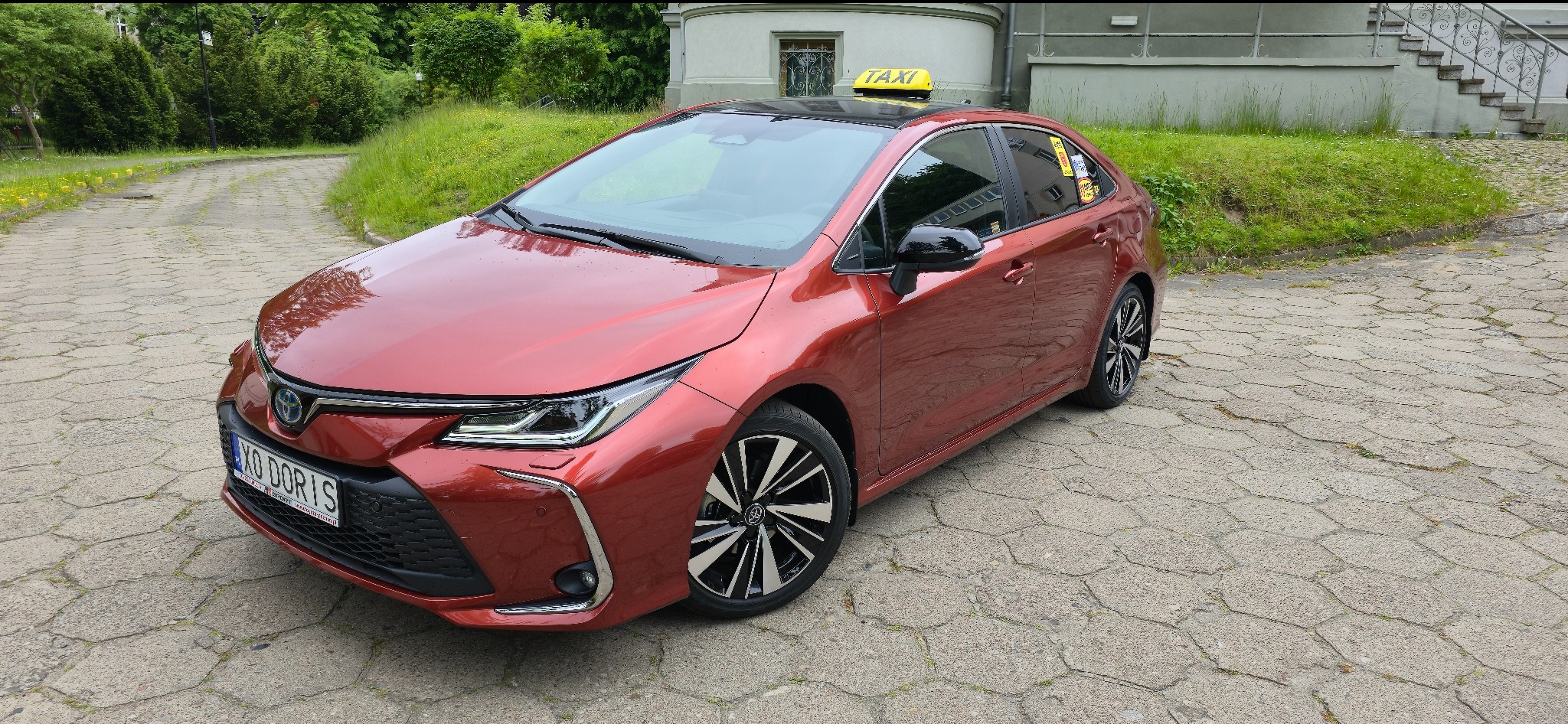 Czerwona Toyota Corolla z żółtym szyldem TAXI na dachu, zaparkowana na brukowanym podjeździe, zieleń w tle. Komfortowy przewóz osób.