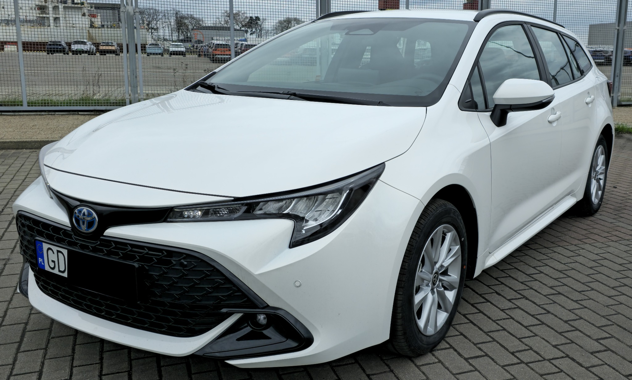 Biała Toyota Corolla kombi, widok z przodu na tle szarej kostki brukowej i metalowego ogrodzenia. Widoczne detale: reflektory, grill, felgi.