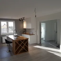 Jasne wnętrze domu po remoncie: białe ściany, drewniany blat wyspy kuchennej z miejscem na wino, szare płytki podłogowe, widoczne ślady po pracach wykończeniowych.