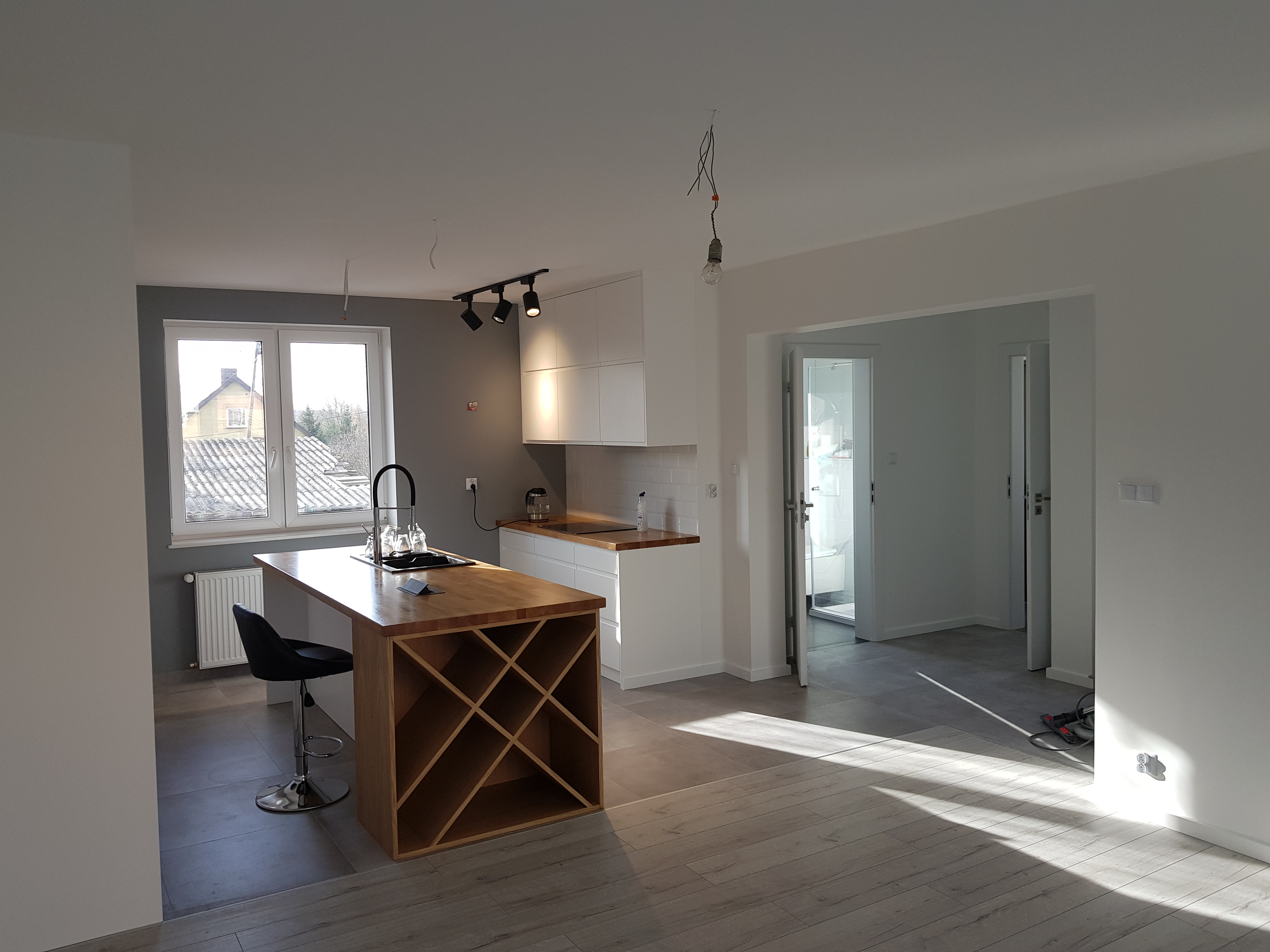 Jasne wnętrze domu po remoncie: białe ściany, drewniany blat wyspy kuchennej z miejscem na wino, szare płytki podłogowe, widoczne ślady po pracach wykończeniowych.