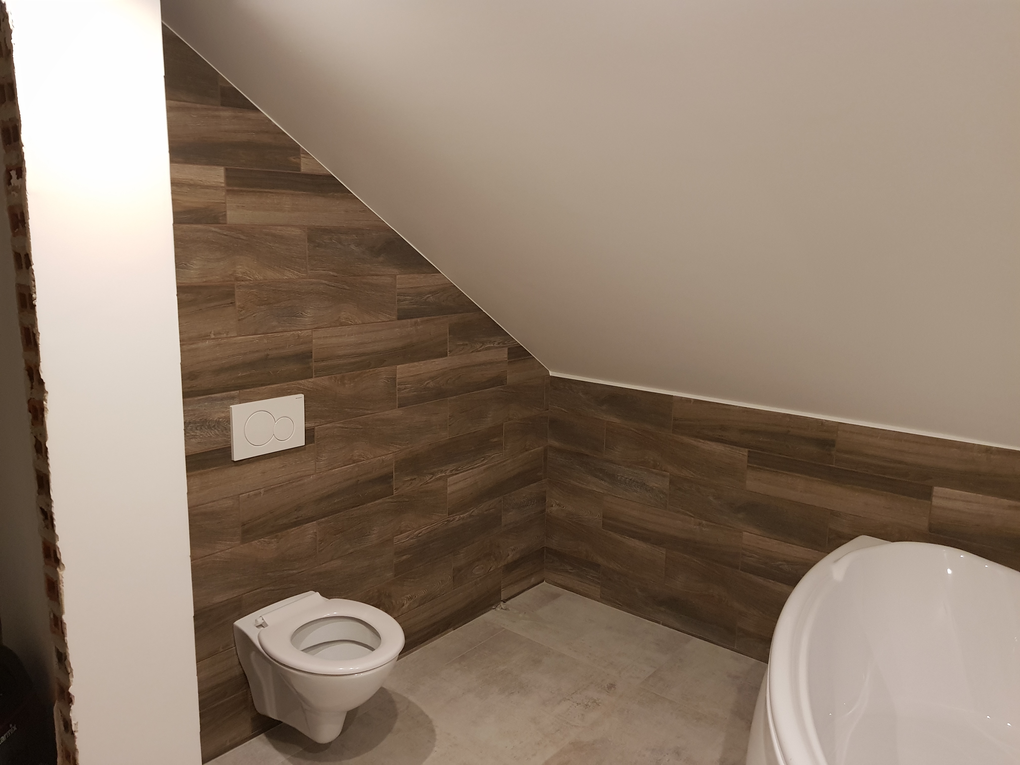 Wykończona łazienka na poddaszu z brązowymi płytkami imitującymi drewno na ścianach, białą podwieszaną toaletą z przyciskiem dwudzielnym i fragmentem białej wanny.