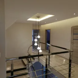 Nowoczesne schody z metalową balustradą i drewnianymi stopniami, oświetlone wiszącą lampą z kilkoma kulistymi kloszami, widok z góry na jasne wnętrze z otwartymi drzwiami.