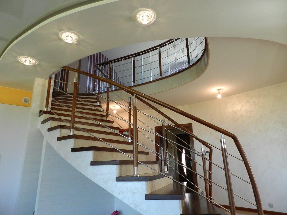 Spiralne schody z drewnianymi stopniami i metalową balustradą w jasnym wnętrzu z podwieszanym sufitem i okrągłymi lampami.