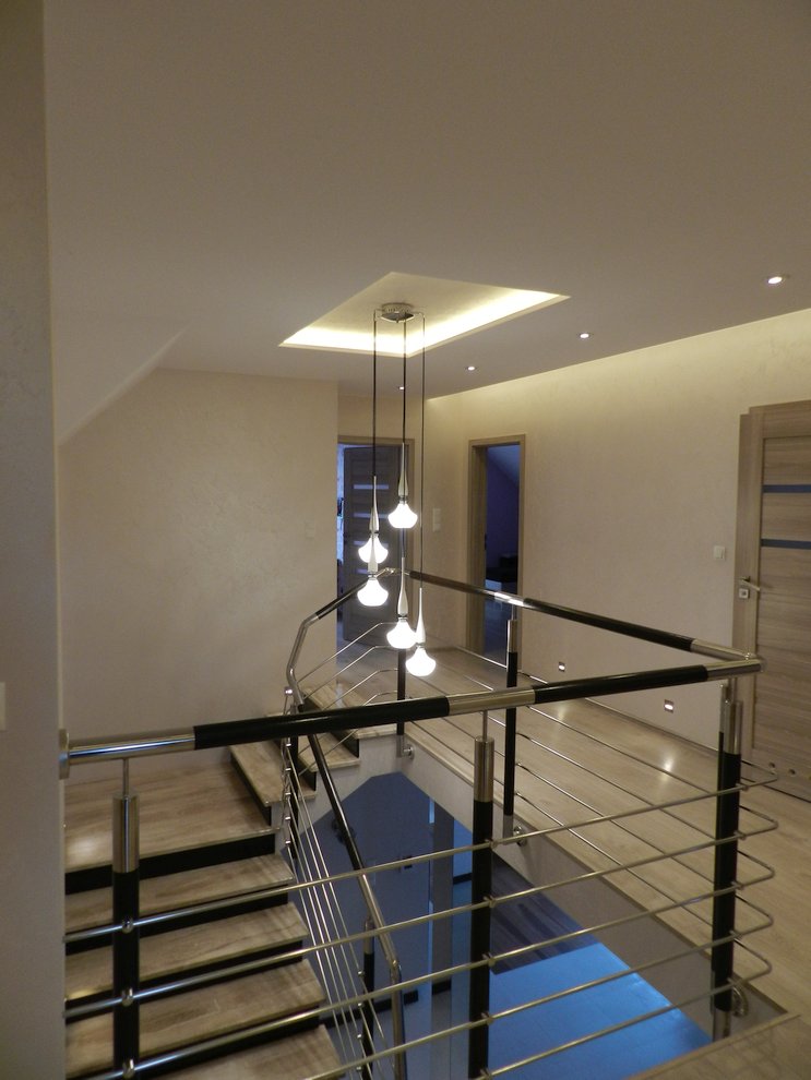 Nowoczesne schody z metalową balustradą i drewnianymi stopniami, oświetlone wiszącą lampą z kilkoma kulistymi kloszami, widok z góry na jasne wnętrze z otwartymi drzwiami.