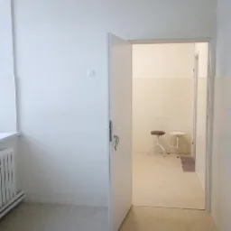 Wnętrze po remoncie: świeżo pomalowane białe ściany, jasna podłoga, otwarte drzwi do pomieszczenia z białymi płytkami i dwoma stołkami.