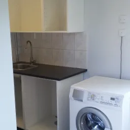 Wnętrze pralni z umywalką w szafce z czarnym blatem, wiszącymi szafkami bez frontów i białą pralką automatyczną.