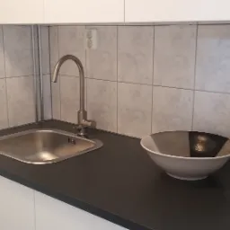 Szare płytki na ścianie nad zlewem ze stali nierdzewnej, ciemny blat i dekoracyjna miska w rogu, widoczne gniazdko elektryczne i włącznik światła.