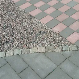 Kompozycja nawierzchni z szarej kostki brukowej ułożonej w jodełkę, różowo-szarej kostki w szachownicę i fragmentu wysypanego drobnym żwirem oddzielonym obrzeżem z kostki.