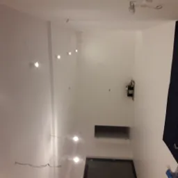 Białe ściany w trakcie wykończenia wnętrza, widoczne punkty świetlne, okno i wystające przewody elektryczne. W tle fragment zasłony.