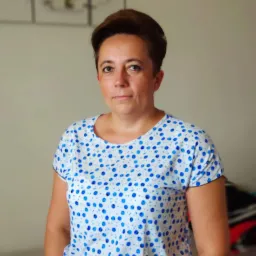 Portret kobiety z kr&oacute;tkimi brązowymi włosami, ubranej w białą bluzkę w niebieskie kropki, na tle beżowej ściany z obrazem kwiat&oacute;w.