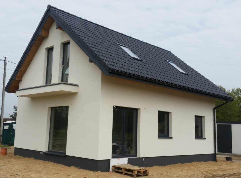 Nowoczesny dom jednorodzinny z jasną elewacją, ciemną dachówką i grafitowymi akcentami, widok z zewnątrz na częściowo zagospodarowany teren.