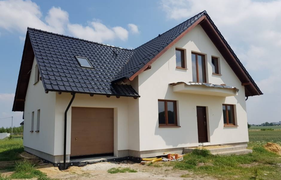 Nowoczesny dom jednorodzinny z jasną elewacją, ciemną dachówką i brązowymi oknami, widoczny z zewnątrz w słoneczny dzień, z elementami wykończeniowymi na balkonie