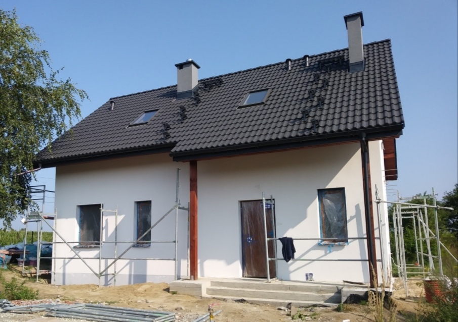 Dwukondygnacyjny dom jednorodzinny z czarną dachówką i białymi ścianami zewnętrznymi w trakcie prac wykończeniowych, widoczne rusztowania i zabezpieczone okna folią, na dachu dwa kominy i okna...
