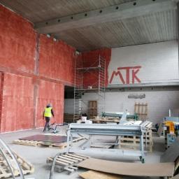 Przedsiębiorstwo MtK - Wnętrze w trakcie prac wykończeniowych: czerwona ściana z nałożoną gładzią, rusztowanie, maszyny do obróbki blachy i pracownik w kamizelce odblaskowej.