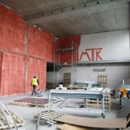 Wnętrze w trakcie prac wykończeniowych: czerwona ściana z nałożoną gładzią, rusztowanie, maszyny do obróbki blachy i pracownik w kamizelce odblaskowej.