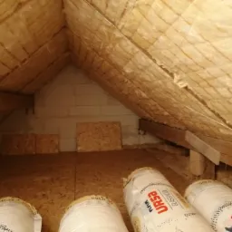 Izolacja poddasza wełną mineralną URSA, widoczne rolki materiału na podłodze z płyt OSB, ocieplony skośny dach i fragment ściany z bloczk&oacute;w.
