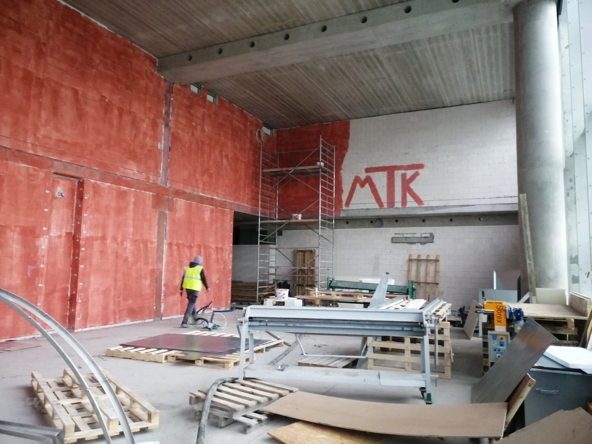 Wnętrze w trakcie prac wykończeniowych: czerwona ściana z nałożoną gładzią, rusztowanie, maszyny do obróbki blachy i pracownik w kamizelce odblaskowej.