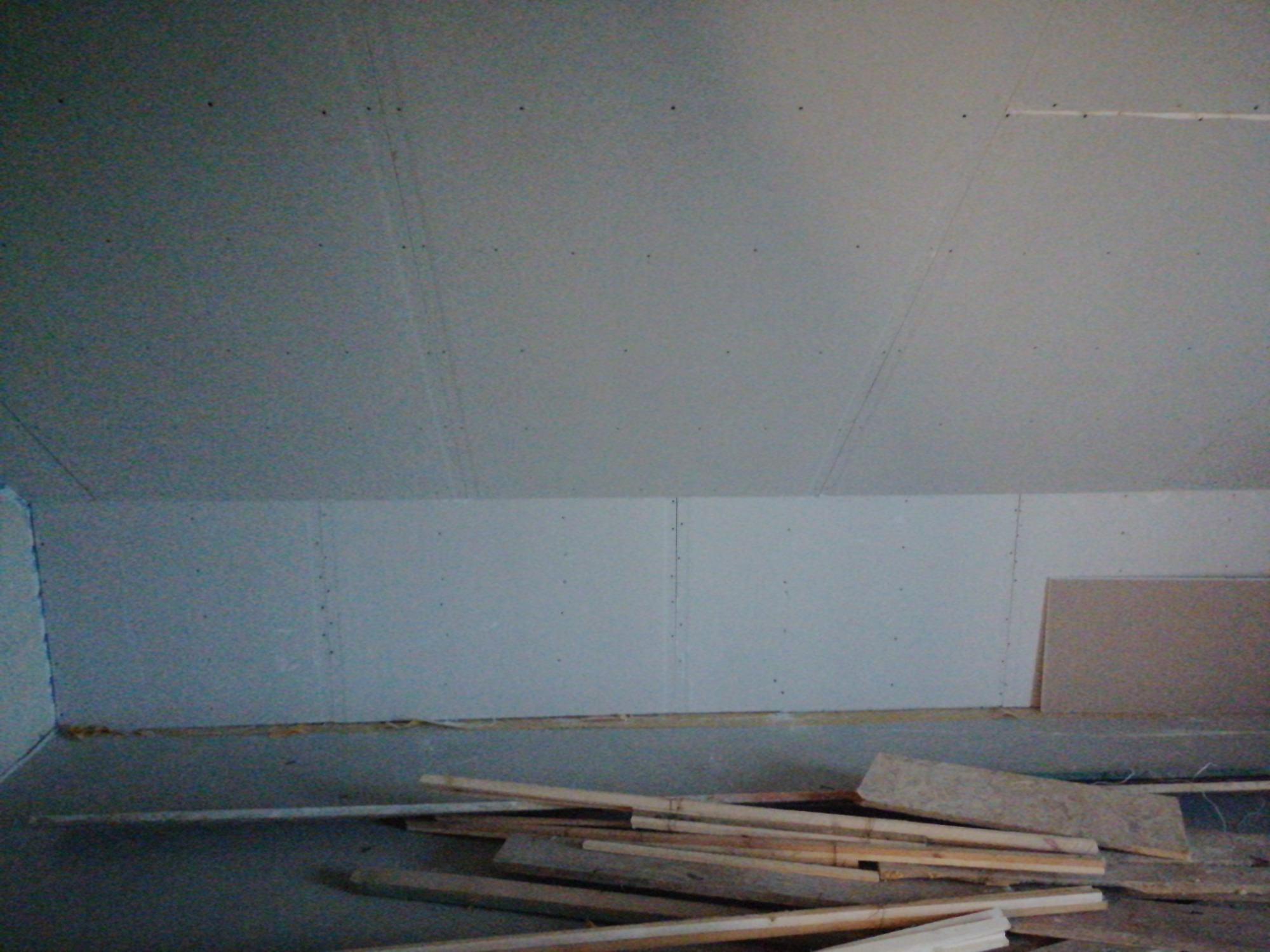 Płyty kartonowo-gipsowe zamontowane na ścianie poddasza, widoczne łączenia i ślady po wkrętach, na podłodze leżą drewniane listwy i fragmenty płyt.