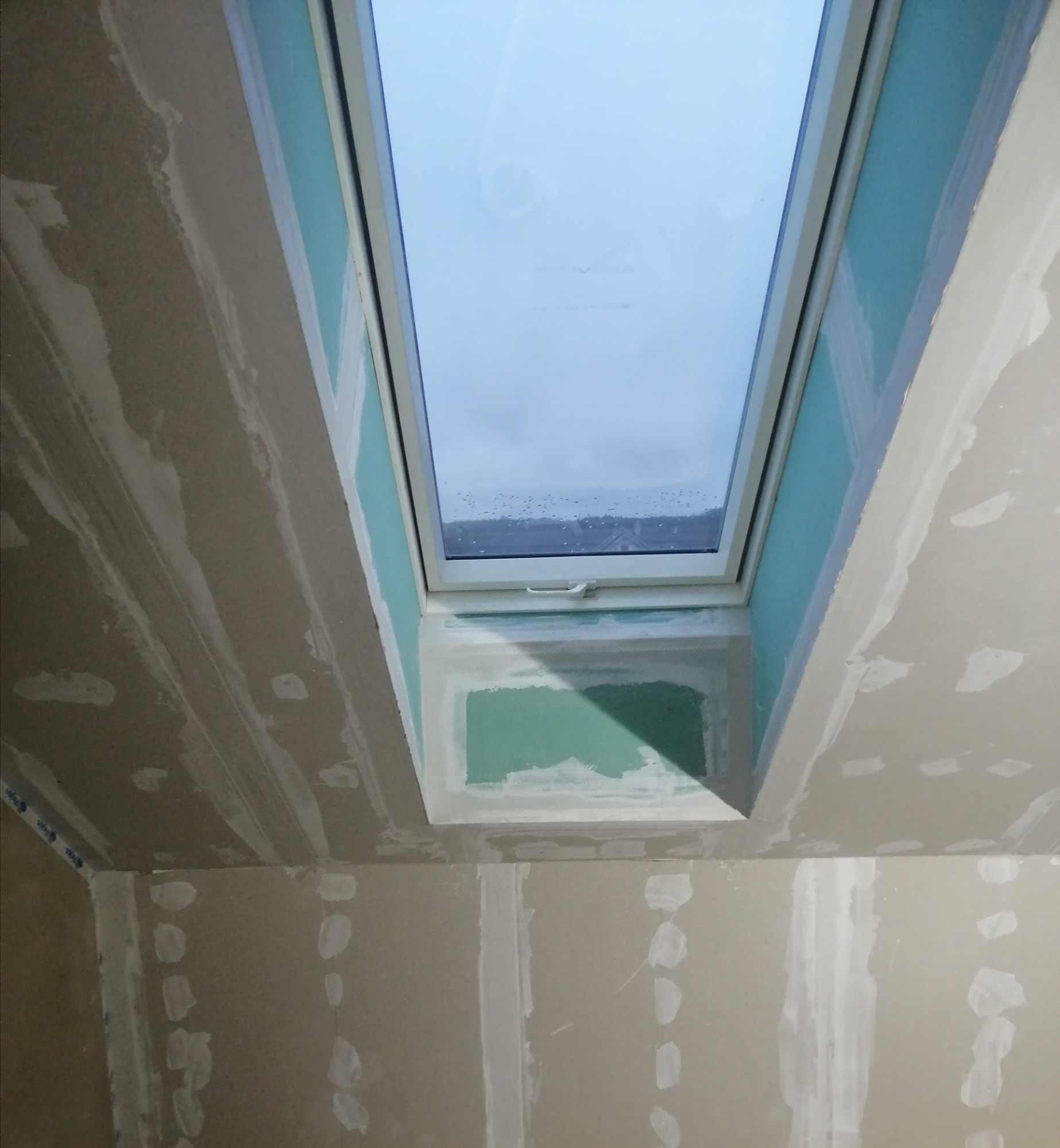 Montaż okna dachowego w surowym wnętrzu, widoczne płyty gipsowo-kartonowe z nałożoną gładzią szpachlową, przygotowane do dalszych prac wykończeniowych.