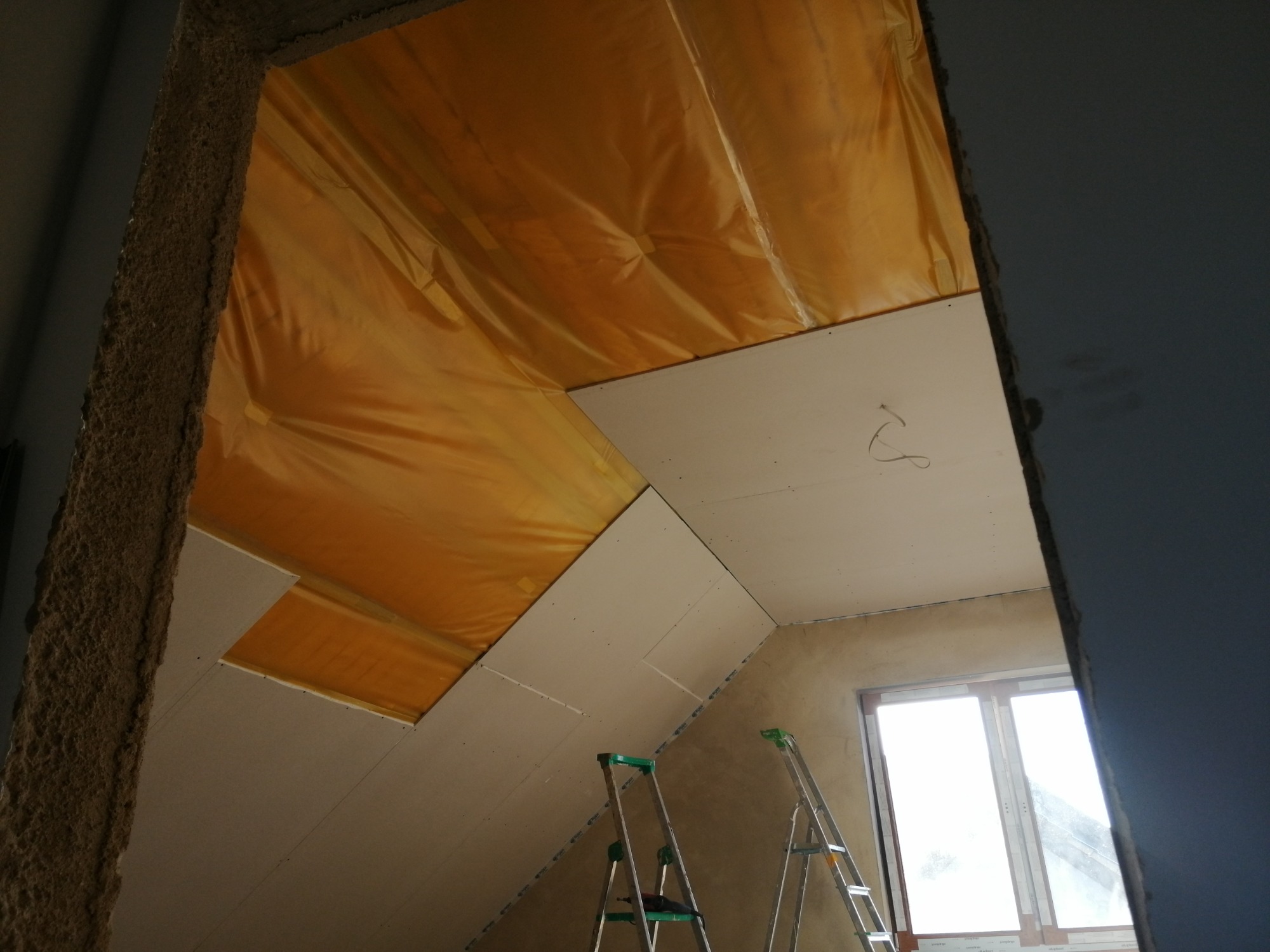 Poddasze w trakcie remontu: widok na skośny sufit z zamontowanymi płytami gipsowo-kartonowymi i folią izolacyjną, dwie drabiny aluminiowe w tle, surowy stan ścian