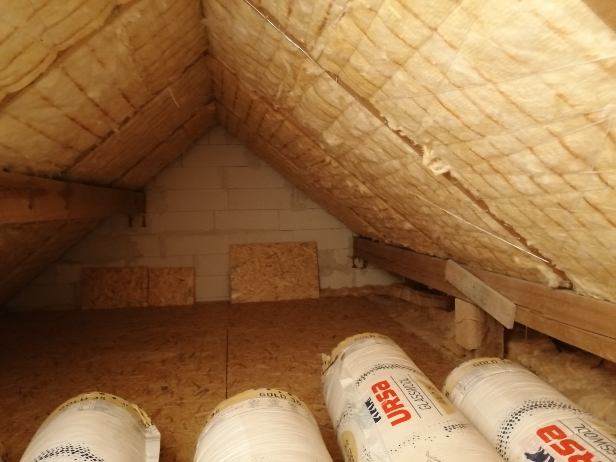 Izolacja poddasza wełną mineralną URSA, widoczne rolki materiału na podłodze z płyt OSB, ocieplony skośny dach i fragment ściany z bloczków.