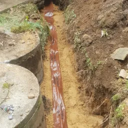 Wykop z ułożoną rurą kanalizacyjną w folii ochronnej, widoczne betonowe kręgi szamba i dwóch pracowników w tle.