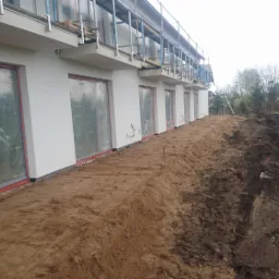 Przygotowany teren pod budowę, równa, piaszczysta powierzchnia wzdłuż ściany budynku z osłoniętymi oknami, widoczne ślady po pracach ziemnych na dalszym planie.