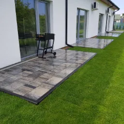 Wyłożony kostką taras przydomowy z grillem, widok na trawnik i elewację budynku z klimatyzacją.