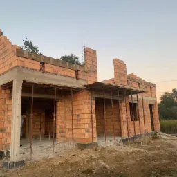 Surowy stan budynku jednorodzinnego z czerwonej cegły, z widocznymi podporami stropu i zbrojeniami, na tle zachodzącego słońca.