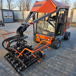 Multi Bruk - Specjalistyczna maszyna H99 do układania kostki brukowej na szarym bruku, z kabiną i ramieniem z chwytakiem. Widoczne detale konstrukcyjne i oświetlenie.