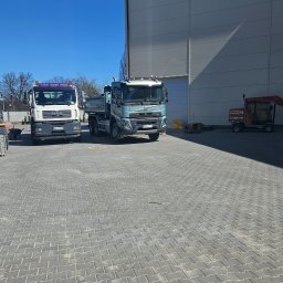 Multi Bruk - Ciężki sprzęt budowlany: koparko-ładowarka z paletą kostki, dwie ciężarówki MAN i Volvo, oraz mała maszyna do układania kostki brukowej na szarym brukowanym placu.