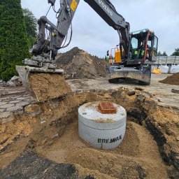 Multi Bruk - Koparka Volvo w trakcie wykopu pod betonowy element studni z napisem 'BREJNAK', widoczne warstwy ziemi i gruzu, w tle hałda ziemi i drzewa.