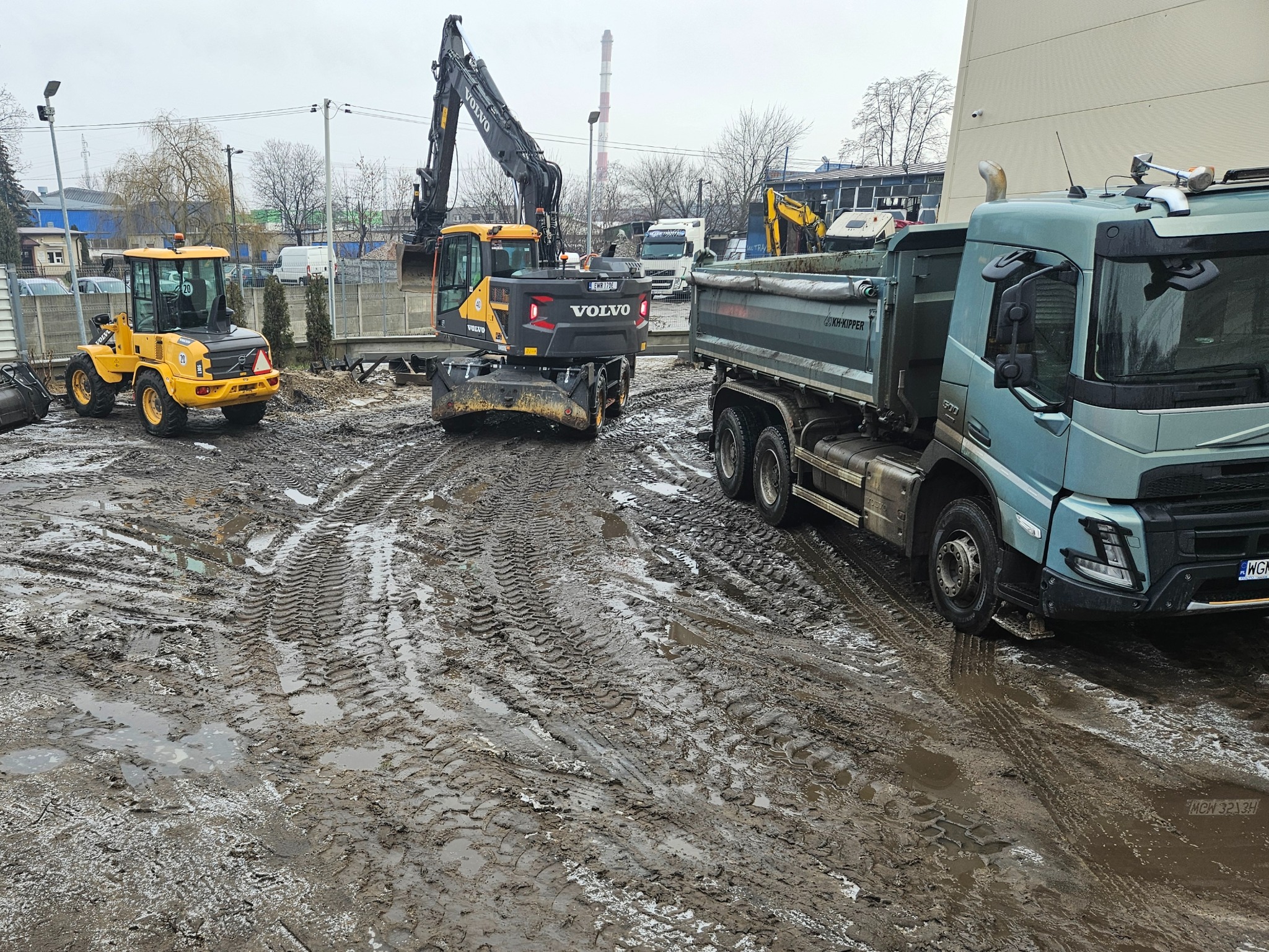 Żółta ładowarka, koparka Volvo i wywrotka na błotnistym placu budowy. Ślady opon w błocie, w tle budynki i komin. Pochmurne niebo.