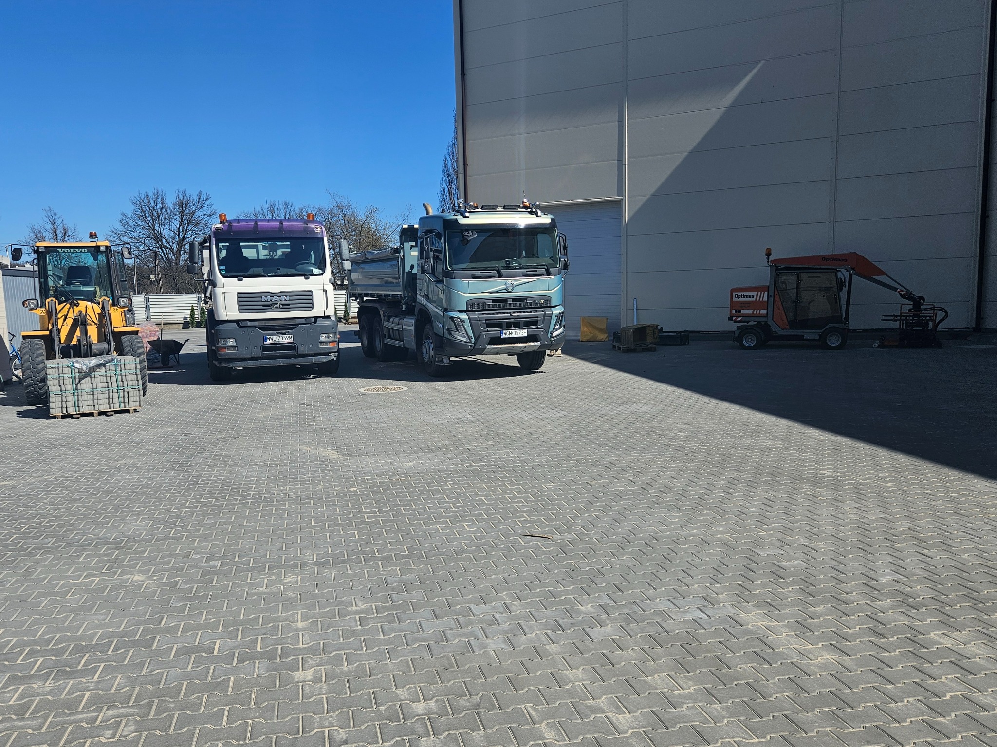 Ciężki sprzęt budowlany: koparko-ładowarka z paletą kostki, dwie ciężarówki MAN i Volvo, oraz mała maszyna do układania kostki brukowej na szarym brukowanym placu.