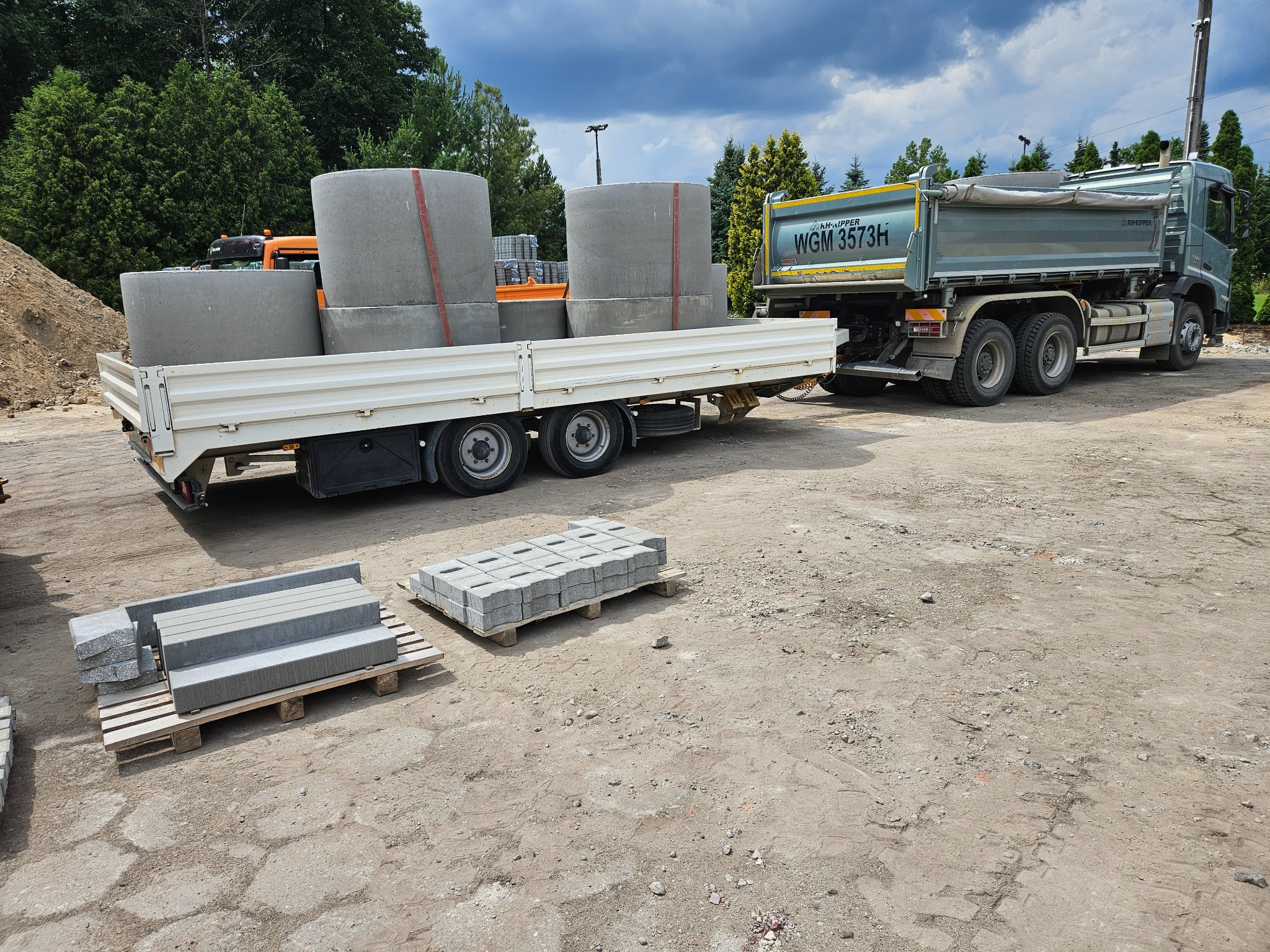 Ciężarówka z przyczepą przewozi betonowe kręgi i kostkę brukową na paletach. W tle drzewa i pochmurne niebo. Widoczny teren budowy.