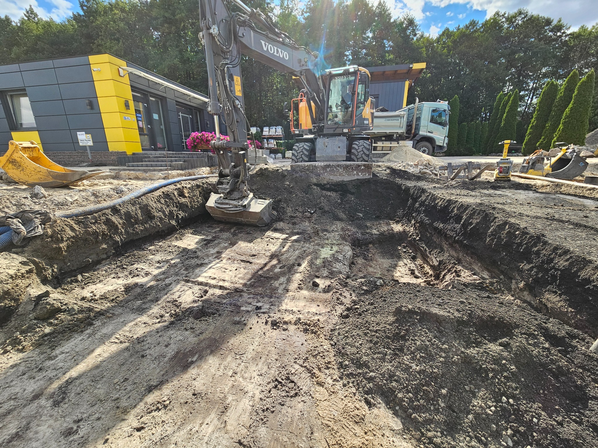 Żółto-szara koparka Volvo wykonuje wykop pod fundamenty na placu budowy. W tle budynek biurowy, ciężarówka i sprzęt budowlany. Słoneczny dzień.