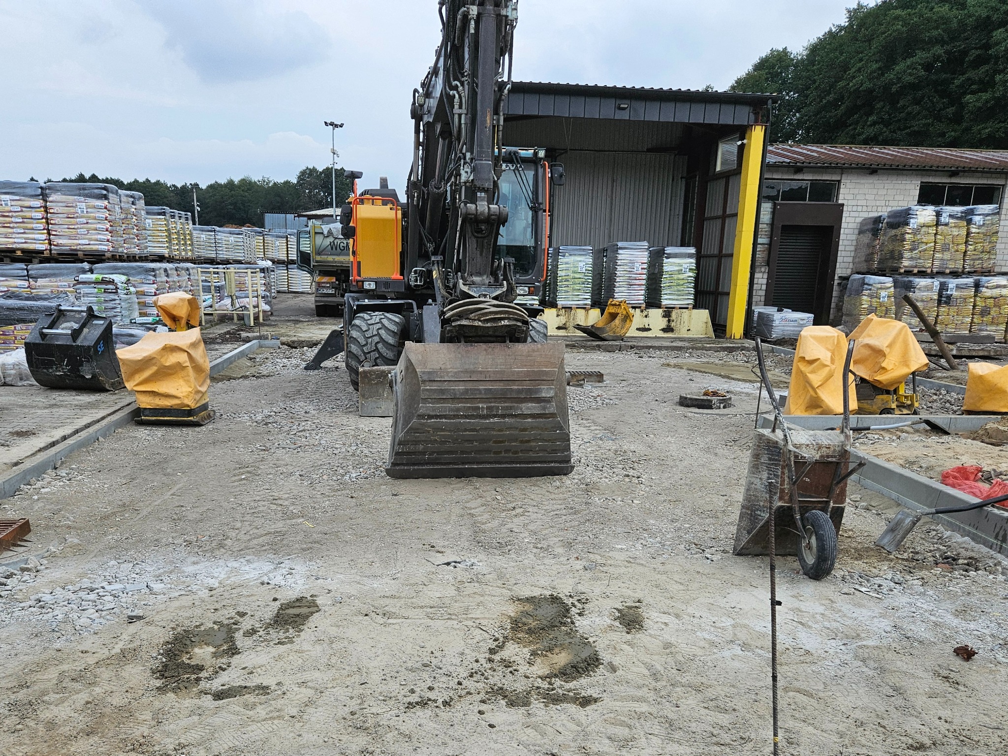 Ciężki sprzęt budowlany, w tym koparka, na placu budowy z materiałami budowlanymi w tle. Widoczne ślady pracy i przygotowany teren pod dalsze prace.