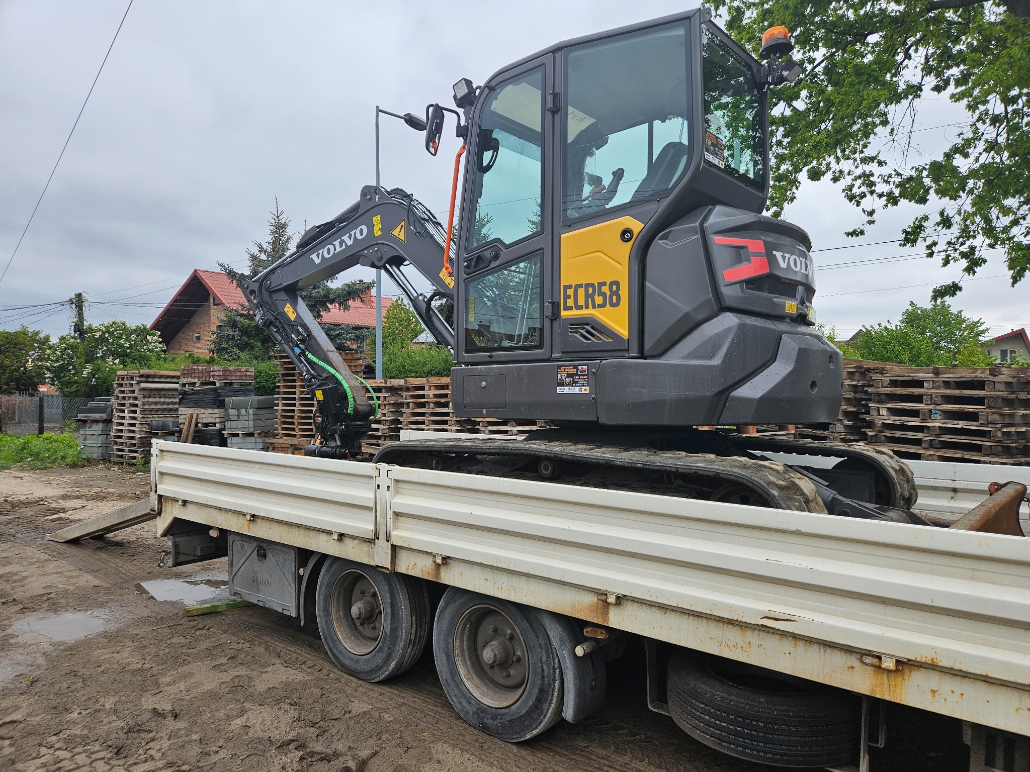Żółto-szara koparka Volvo ECR58 na przyczepie transportowej, z paletami kostki w tle. Widoczne elementy otoczenia: dom z czerwoną dachówką i zieleń.