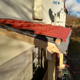 Montaż czerwonej blachodachówki na drewnianej konstrukcji dachu, widoczne narzędzia i materiały budowlane, w tle ściana budynku z oknem i anteną satelitarną.