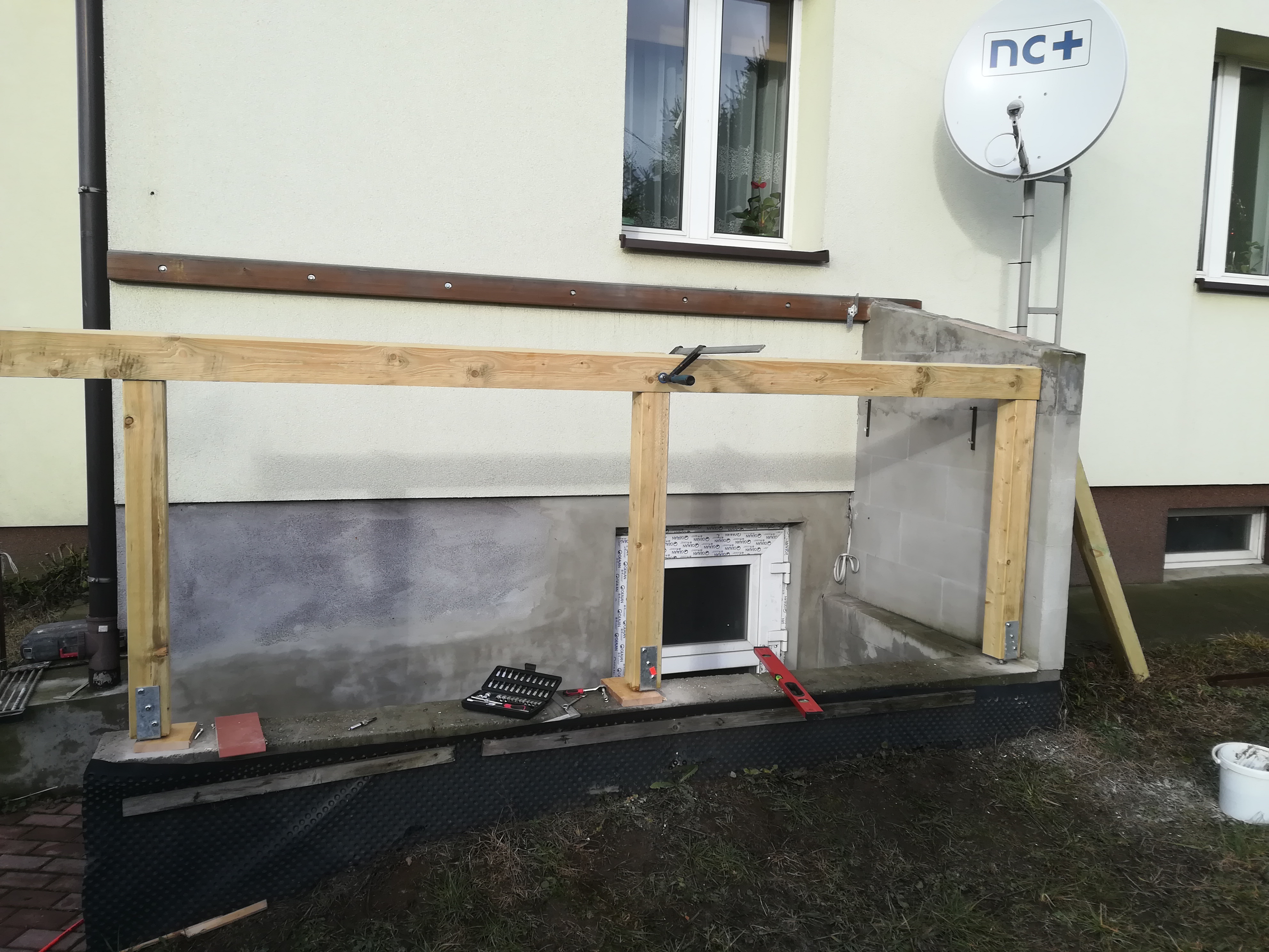 Drewniana konstrukcja w trakcie budowy przy ścianie domu z oknem i anteną satelitarną. Widoczne narzędzia, poziomica i skrzynka z narzędziami.