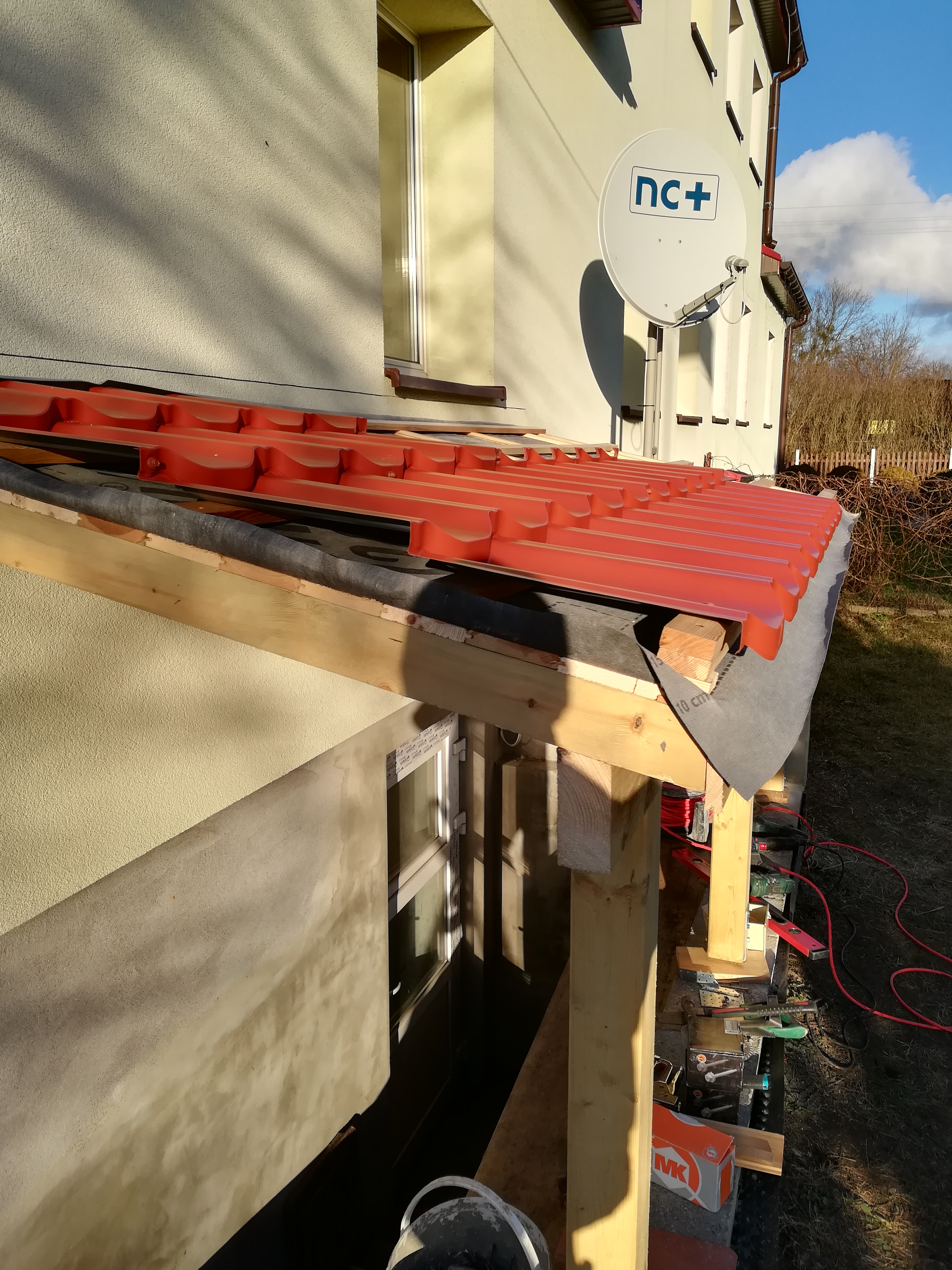 Montaż czerwonej blachodachówki na drewnianej konstrukcji dachu, widoczne narzędzia i materiały budowlane, w tle ściana budynku z oknem i anteną satelitarną.