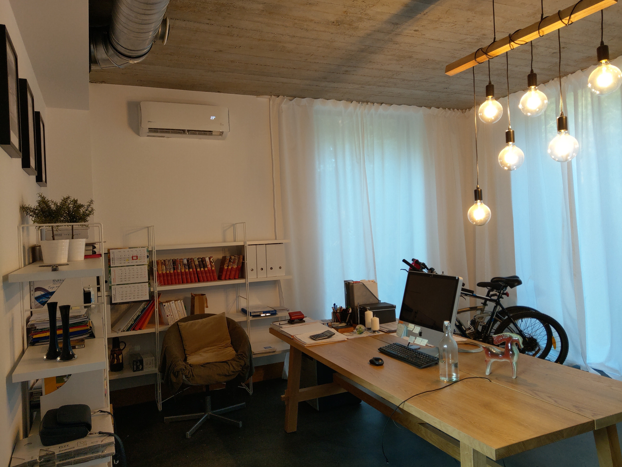 Wnętrze jasnego biura domowego z klimatyzatorem ściennym, drewnianym biurkiem, komputerem, rowerem i dekoracyjnymi lampami wiszącymi.