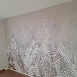 Ściana w pokoju pomalowana farbą w odcieniach szarości z motywem białych liści palmowych, widoczna goła żarówka na kablu.