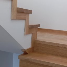 Well Done - Drewniane schody o jasnym odcieniu dębu, z widocznymi detalami łączeń i minimalistycznym wykończeniem, osadzone w białym wnętrzu z delikatnym oświetleniem.