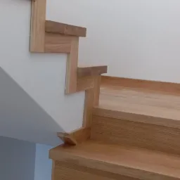 Drewniane schody o jasnym odcieniu dębu, z widocznymi detalami łączeń i minimalistycznym wykończeniem, osadzone w białym wnętrzu z delikatnym oświetleniem.