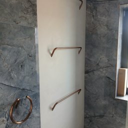 Well Done - Wykończona łazienka: szare marmurowe płytki, biała ściana z trzema miedzianymi wieszakami na ręczniki i okrągły wieszak obok lustra. Minimalistyczny design.